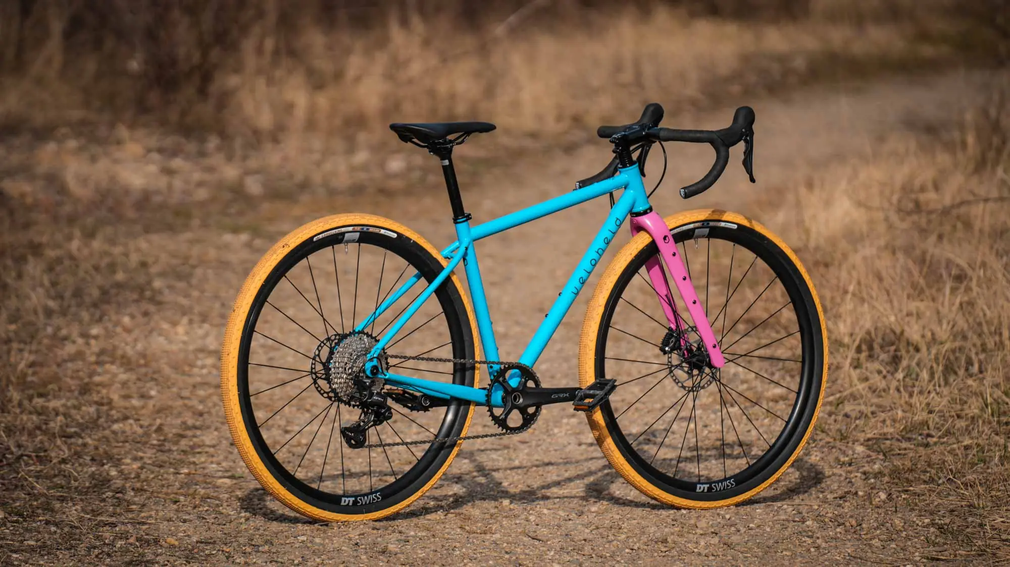 veloheld iconX Custom Gravelbike Bubblegum Farbdesign Detail mit blauem Rahmen und pinker Gabel und gelben Reifen - Komplettrad auf Schotter stehend