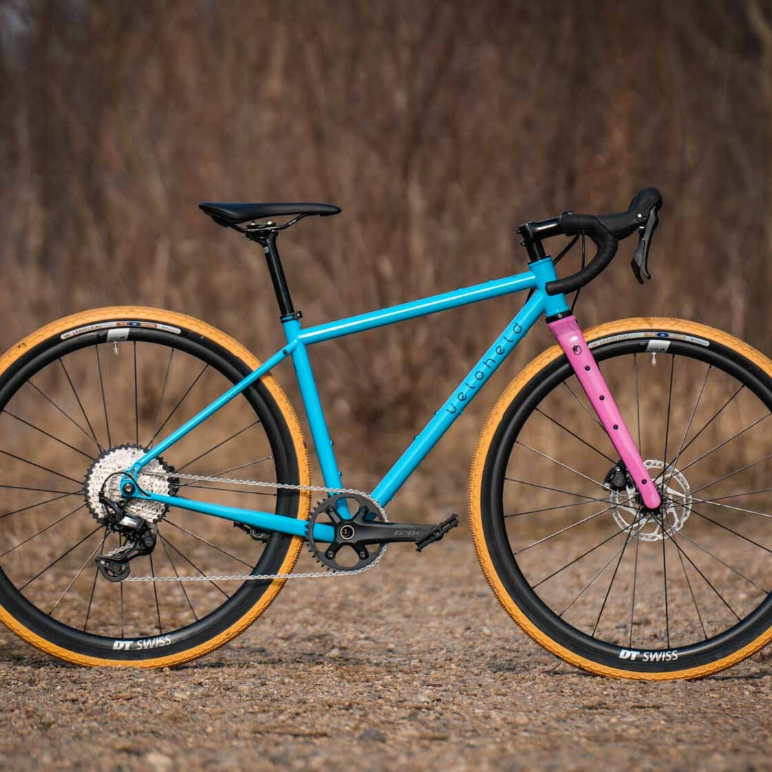 veloheld iconX Custom Gravelbike Bubblegum Farbdesign Detail mit blauem Rahmen und pinker Gabel und gelben Reifen - Komplettrad auf Schotter stehend