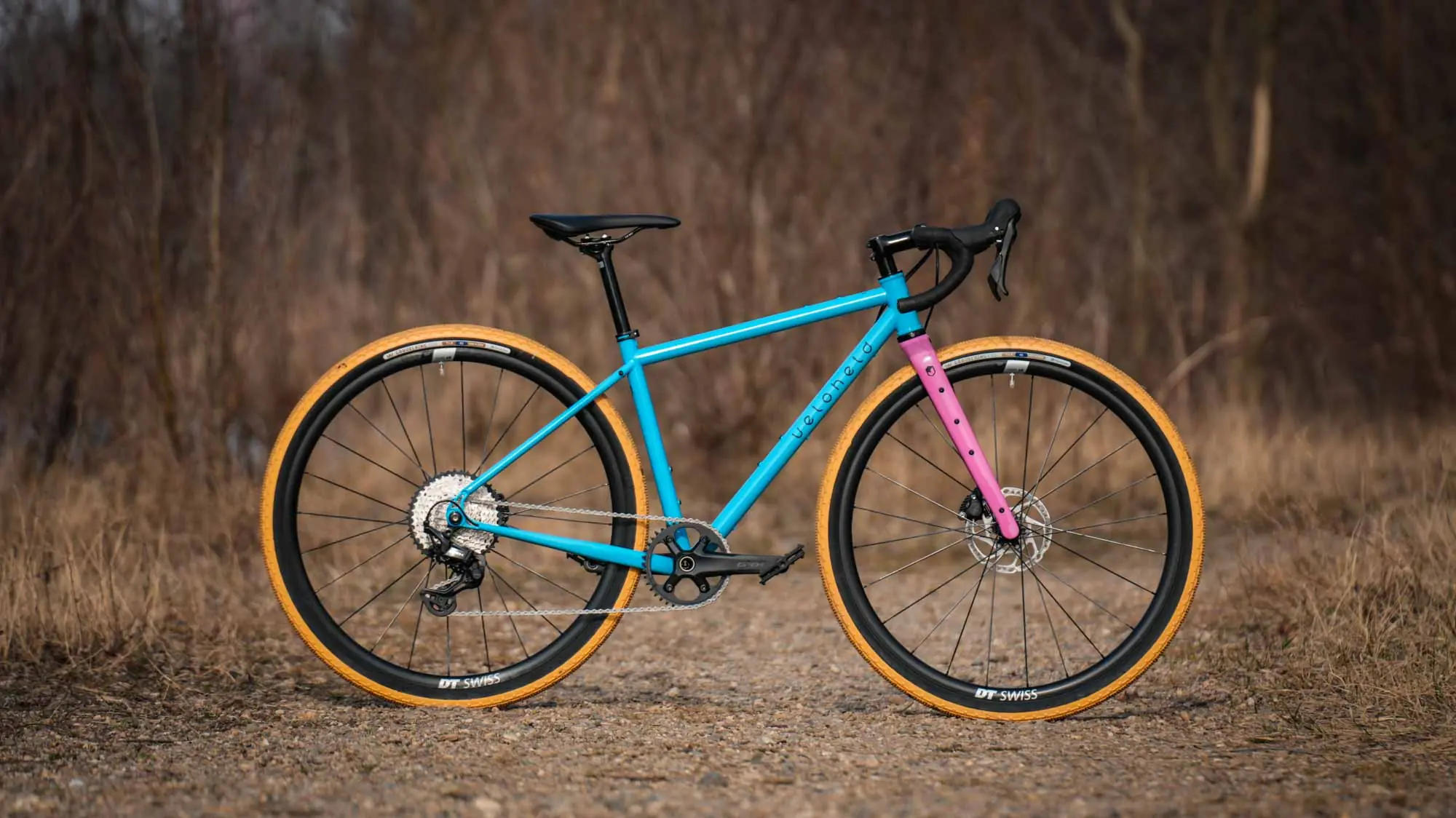 veloheld iconX Custom Gravelbike Bubblegum Farbdesign Detail mit blauem Rahmen und pinker Gabel und gelben Reifen - Komplettrad auf Schotter stehend