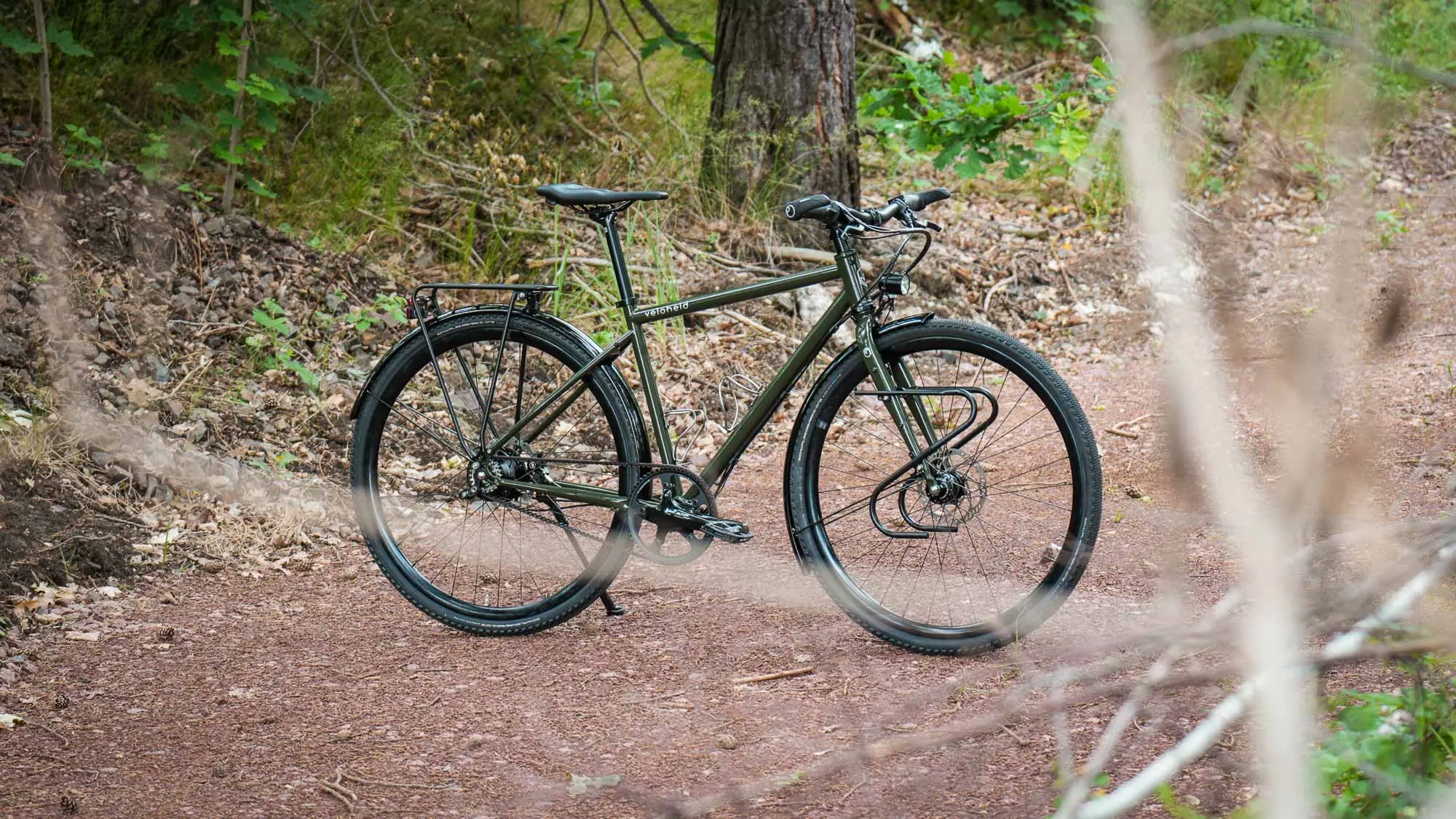 Grünes veloheld.alleyX mit Rohloff-Nabenschaltung, Gates-Riemenantrieb, Schutzblechen und Frontgepäckträger auf einem Waldweg zwischen Bäumen und natürlicher Umgebung.